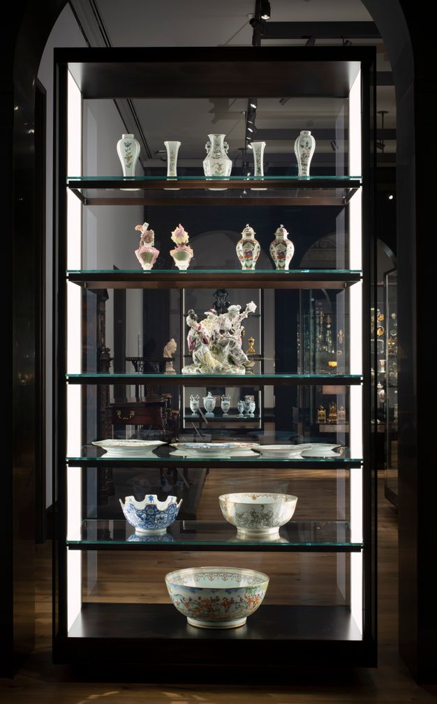 Installation. Six shelves hold porcelains in a case with glass on both long sides so we look through it. From the bottom up, the first two shelves hold bowls, and then a shelf has several plates. A sculptural piece is next, below two shelves of vases and urns.