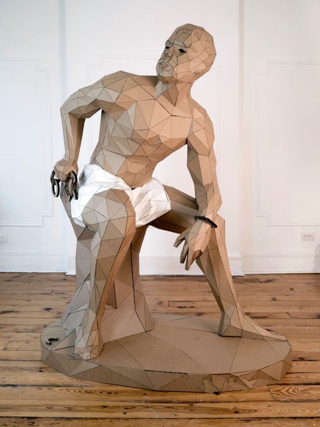 Sculpture. This free-standing sculpture is made of triangular and trapezoidal pieces of cardboard. It shows a man sitting on a stump, leaning forward with one elbow resting a knee. That arm has a shackle on the wrist. His skin, hair, and the base are all the brown of the cardboard. White pieces make a loincloth.