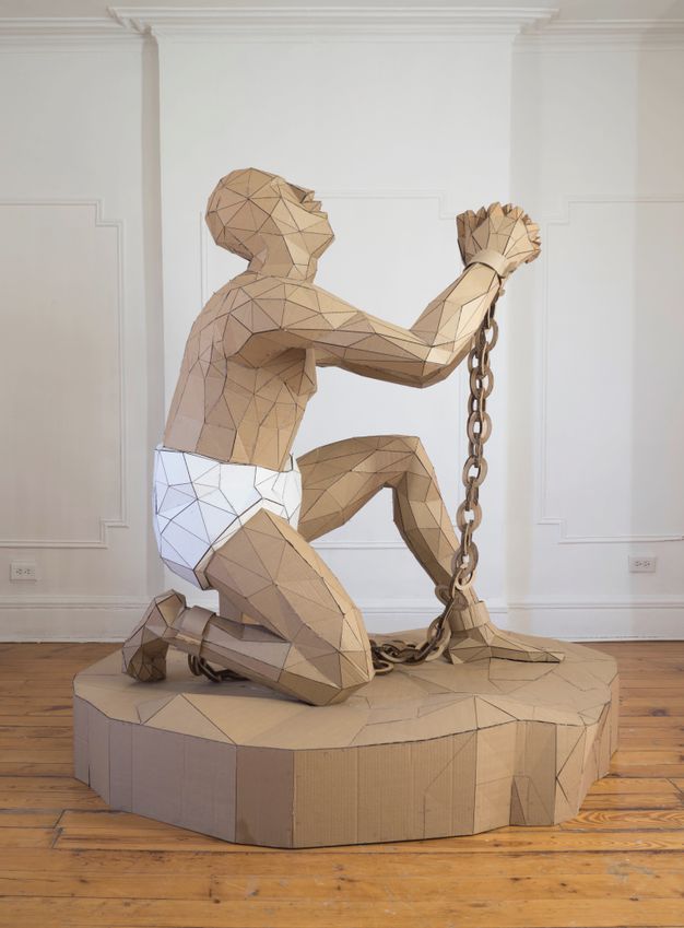 Sculpture. A man made of mostly triangular pieces of cardboard knees on one knee. His other foot is forwards, his hands clasped and raised. Chains connect shackles on his wrists and knees. His head tips back. He faces our right in profile here.