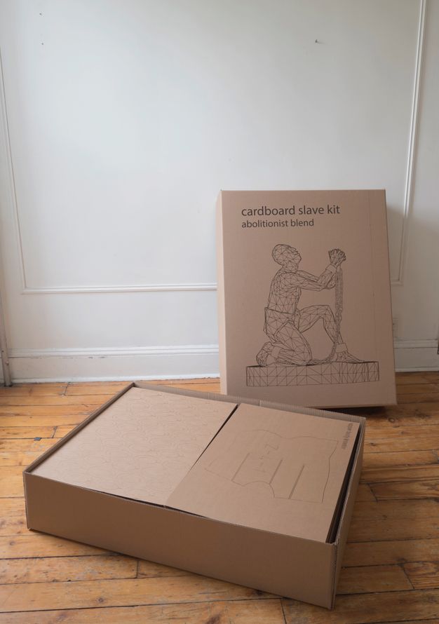 A box holding sheets of cardboard sits on a wood floor in front of the lid, which is propped on the wall behind it. The lid has the title printed above a line drawing of the sculpture the buyer can build with the kit.