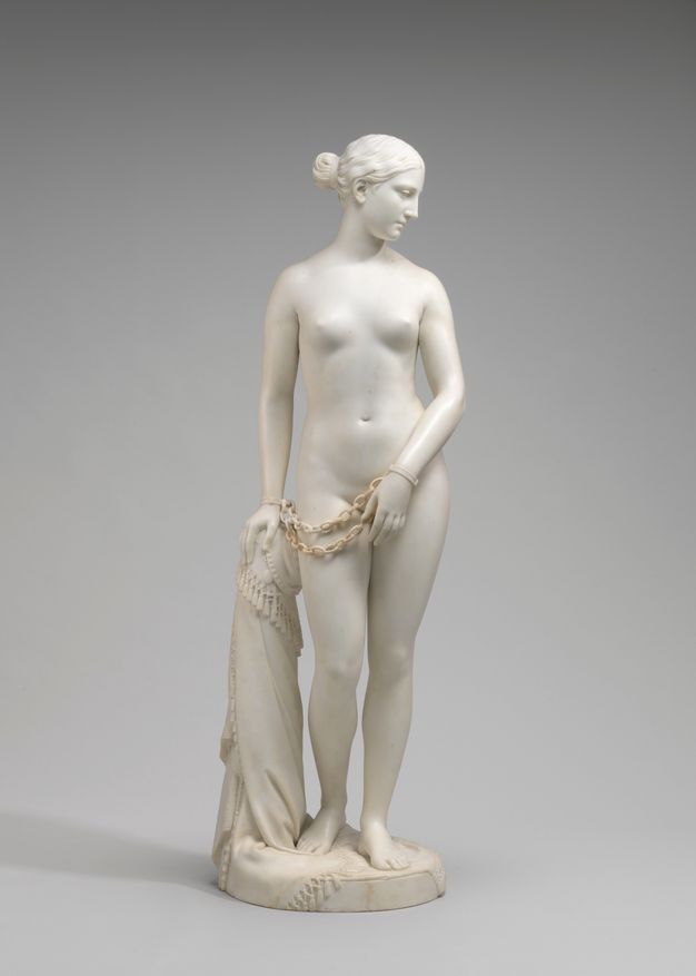 Sculpture. This free-standing statue of a nude woman standing with one hand on a hip-high post, her body facing us as she looks down and to our right in this view. Chains hang from shackles around her wrists.