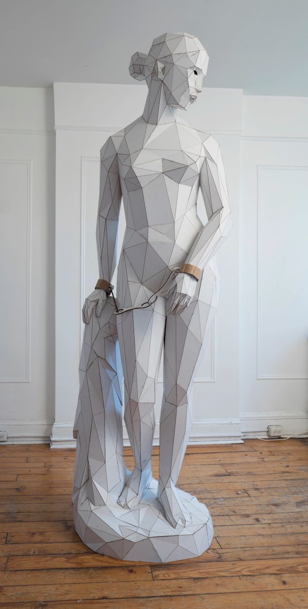 Sculpture. Made with triangular and trapezoidal pieces of cardboard, a nude woman stands and looks over one shoulder, her wrists chained together. The shapes are white except for the manacles, which are the brown of the cardboard.