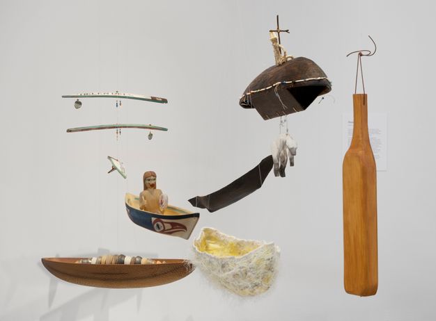 Sculpture. Several canoes and canoe-shaped objects hang against a white gallery wall. One appears to be made from wood and one from a fuzzy, white material. A bottle-shaped plank or paddle hangs to our right.