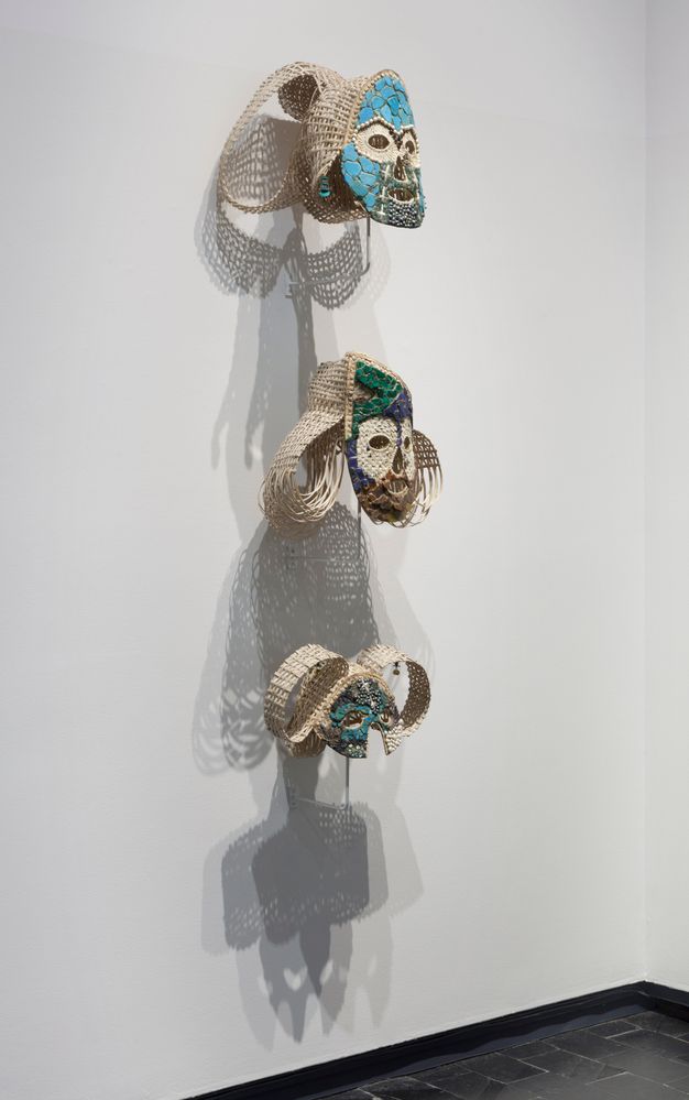 Installation. Three woven masks hang in a column against a white gallery wall. They are made with off white, emerald-green, and turquoise-blue material. Lacy shadows make intricate geometric patterns on the wall behind them.