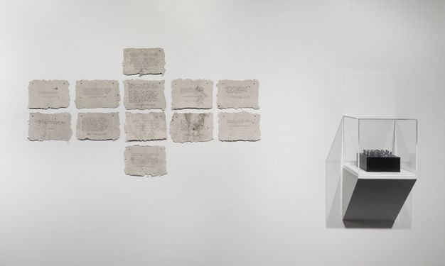 Installation. Two works are hung on a white wall. To our left, twelve rough-edged, rectangular pieces of paper appear nailed or tacked to the wall. A column of four papers down the middle is flanked by a square of four to each side. A black and possibly silver object is in a case to our right.