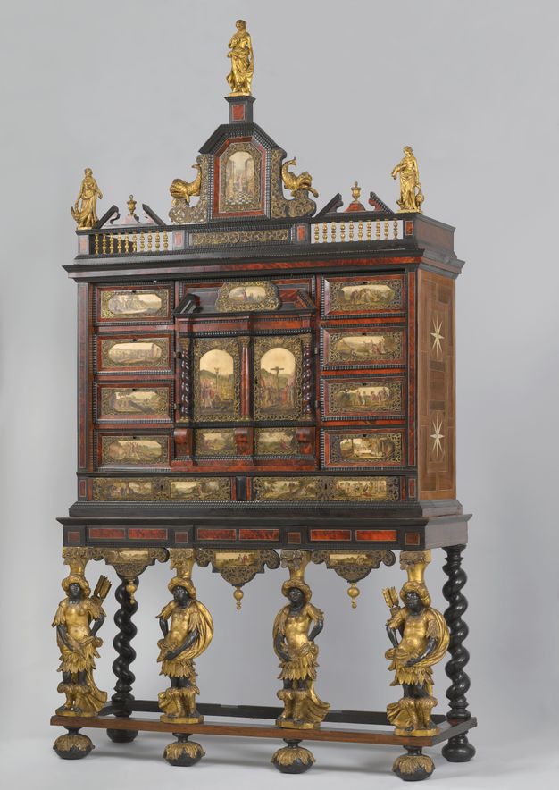 Cabinet. A chest of drawers is topped by a miniature balustrade lined with figures and ornament above. Below, four carved people line the front and act as legs. The drawers and a cabinet at the centre are fronted with paintings on marble. The people have black-coloured skin and wear gold turbans, clothing, and leg coverings.