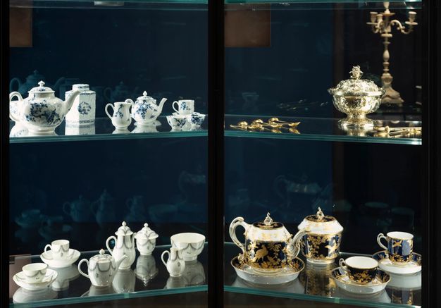 Installation. Four glass shelves in a cabinet hold porcelain, silver, and gold vessels, containers, and utensils for storing and making tea. The objects reflect in the glass on the back side and in the glass of the shelves, creating double and sometimes triple vision.