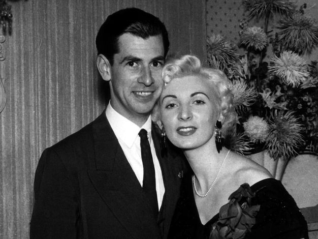 Black and white photograph. A man wearing a suit and a woman in a dark dress and pearls pose, her forehead touching his cheek, in front of a bouquet of spiky flowers. Both have pale skin. The man has dark hair, and the woman’s blond curls frame her heart-shaped face.
