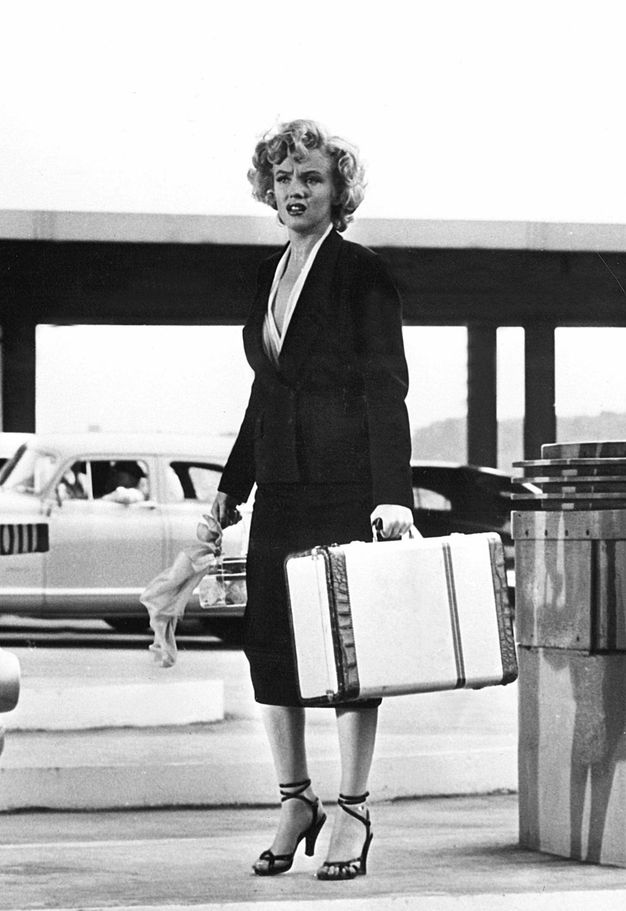 Film still. A pale-skinned woman with blond curly hair stands holding a suitcase and wearing a black skirt suit. Her brows are furrowed as she looks to our left, her full lips parted. Her high-heeled black sandals have straps up the ankles and over the toes.