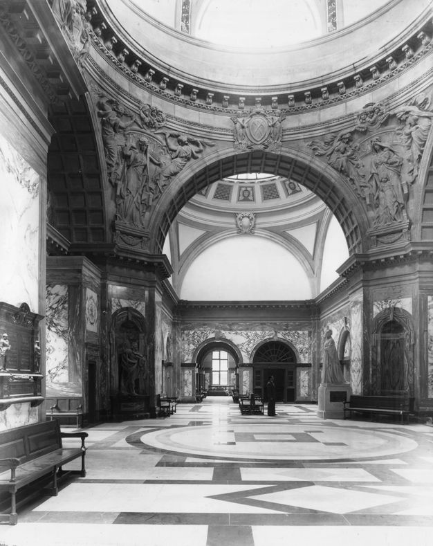 Architecture. Black and white photograph of monumental arches supporting two domes over patterned, tiled floors. Relief carvings in the spandrels we can see are at least twice as tall as a person who stands near a bench across the space from us.