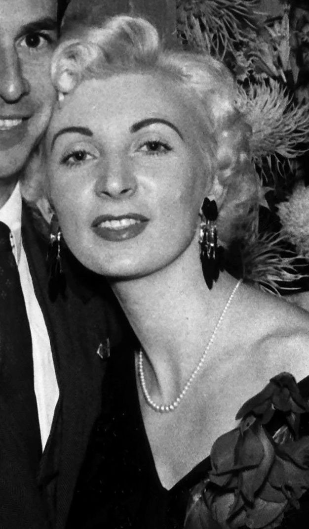 Black and white photograph. Close-view of a pale-skinned woman with shiny blond curls. Her arched eyebrows are dark. Her head slightly tips back. She smiles, and has a full lower lip with a contoured upper lip. Long black earring dangle from her ears, and a thin string of pearls reaches to the hollow of her throat.