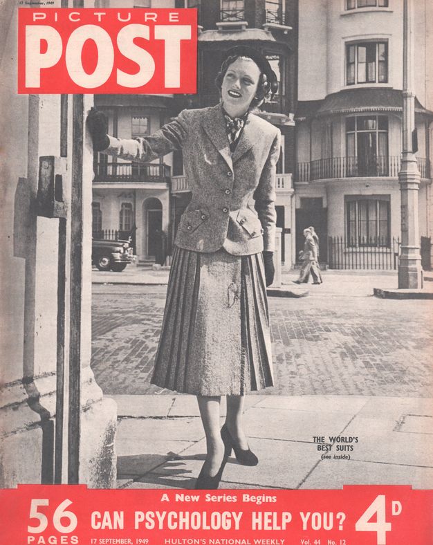 Magazine cover. A pale-skinned woman stands on a street corner, one black-gloved hand against the corner of the building as she peers around and down the street. She wears a fitted, hip-length jacket and a pleated, shin-length skirt. Her black pumps have rounded toes, and she wears a black cap on her curled hair.